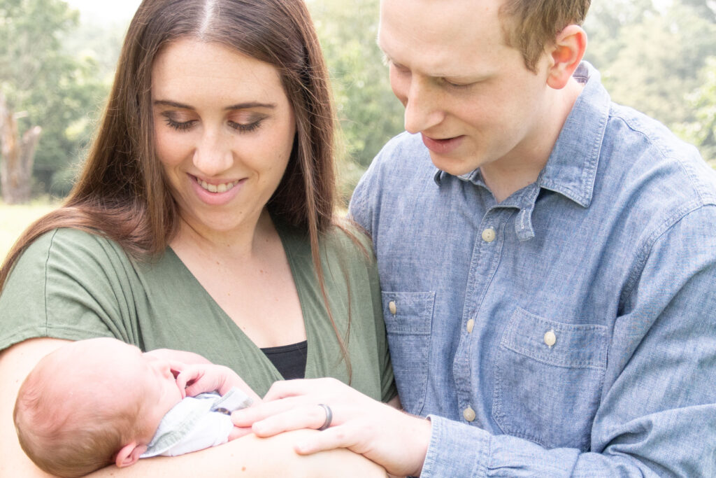 Mom in green dress and dad in blue shirt. They are holding and looking at their newborn baby outside. Landon Lifestyle Newborn session. 