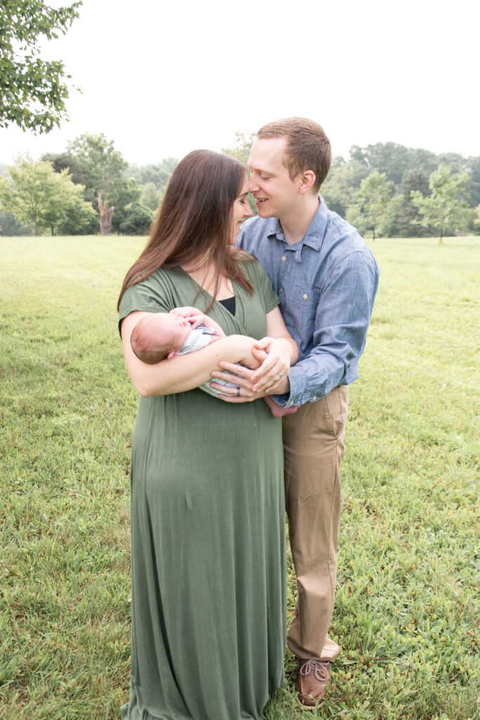 Mom in green dress and dad in blue shirt. They are holding their newborn baby outside.