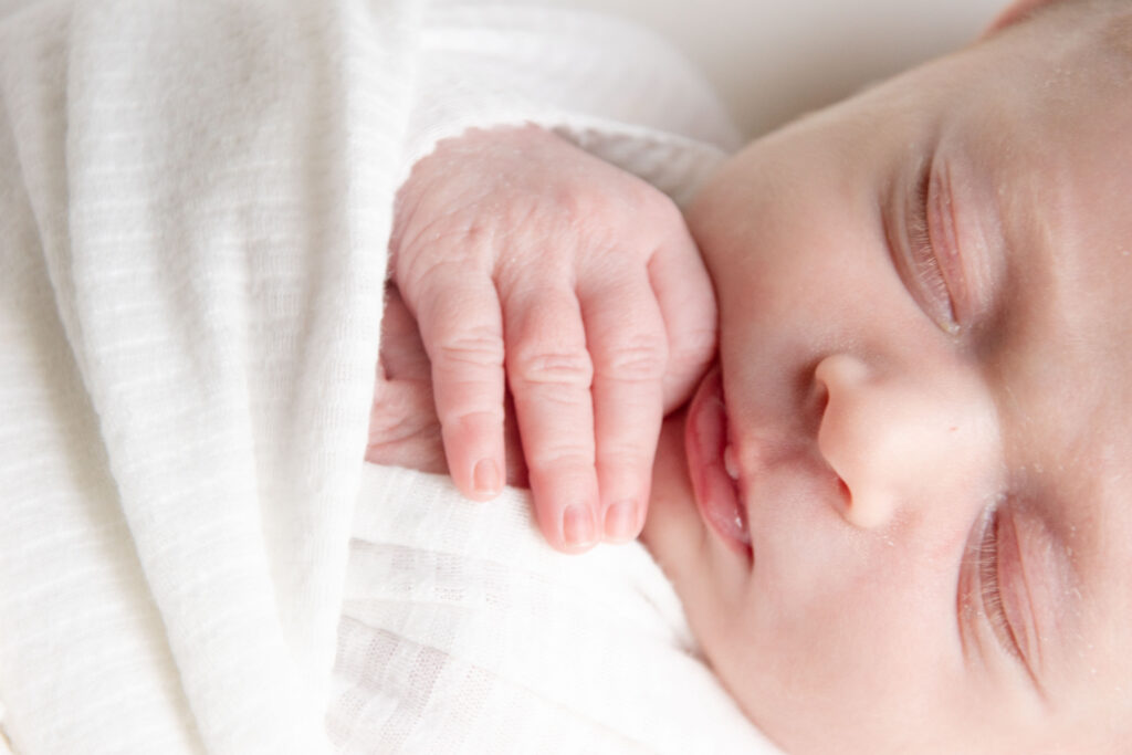 Newborn with hands folded near his face. The baby is wrapped in a white swaddle. Landon Lifestyle Newborn session.