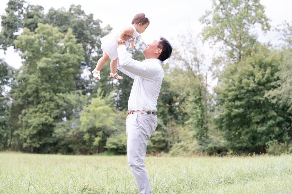 Dad holding toddler in the air wearing white outfits. 