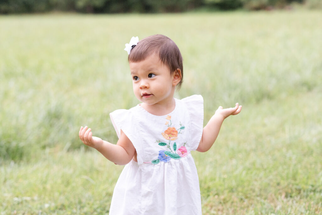 Toddler in white dress with flowers helping her hands out to the side.