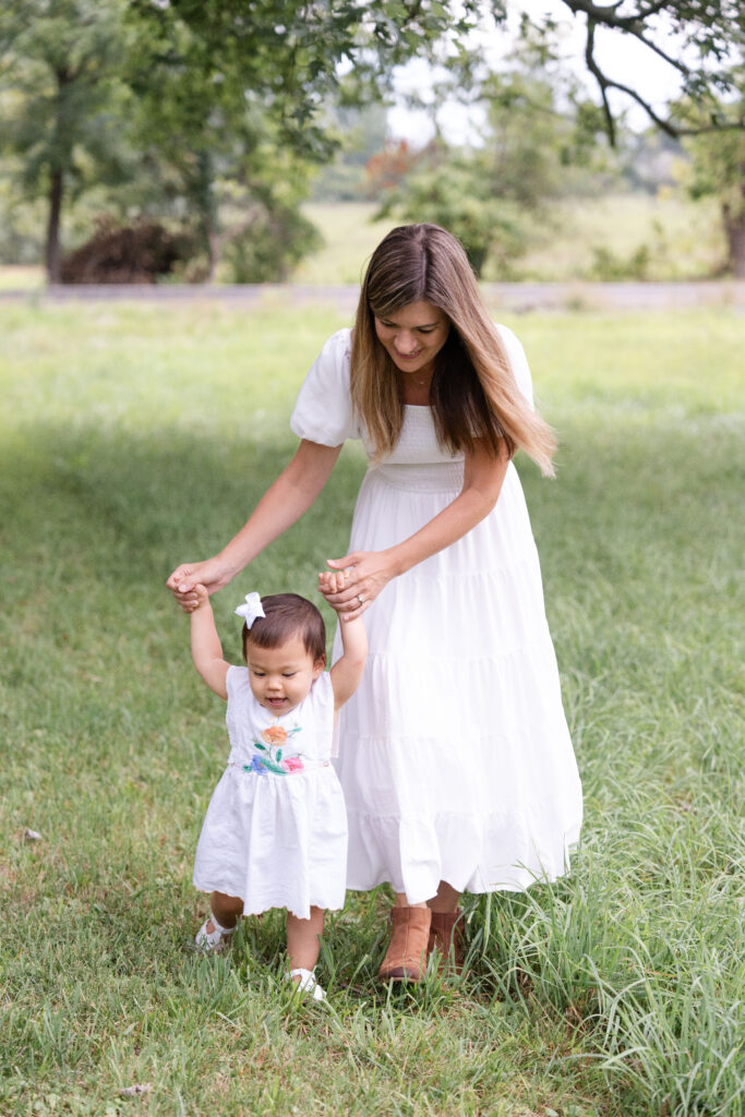 Toddler holding hands being helped by her mom through a field. 