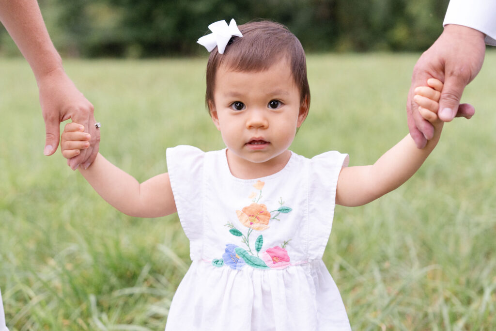 Toddler wearing a white bow holding her parents hands. 